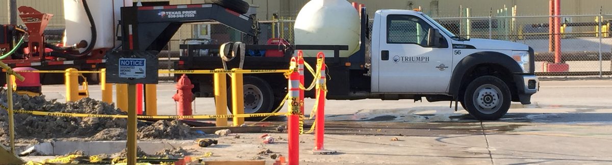 Triumph truck and vacuum tank at a barricaded excavation, with caution tape, cones and bollards protecting the work area